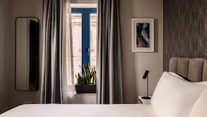 Habitación de hotel con cama, mesita de noche, lámpara, cuadro enmarco, ventana con cortinas y planta en maceta.