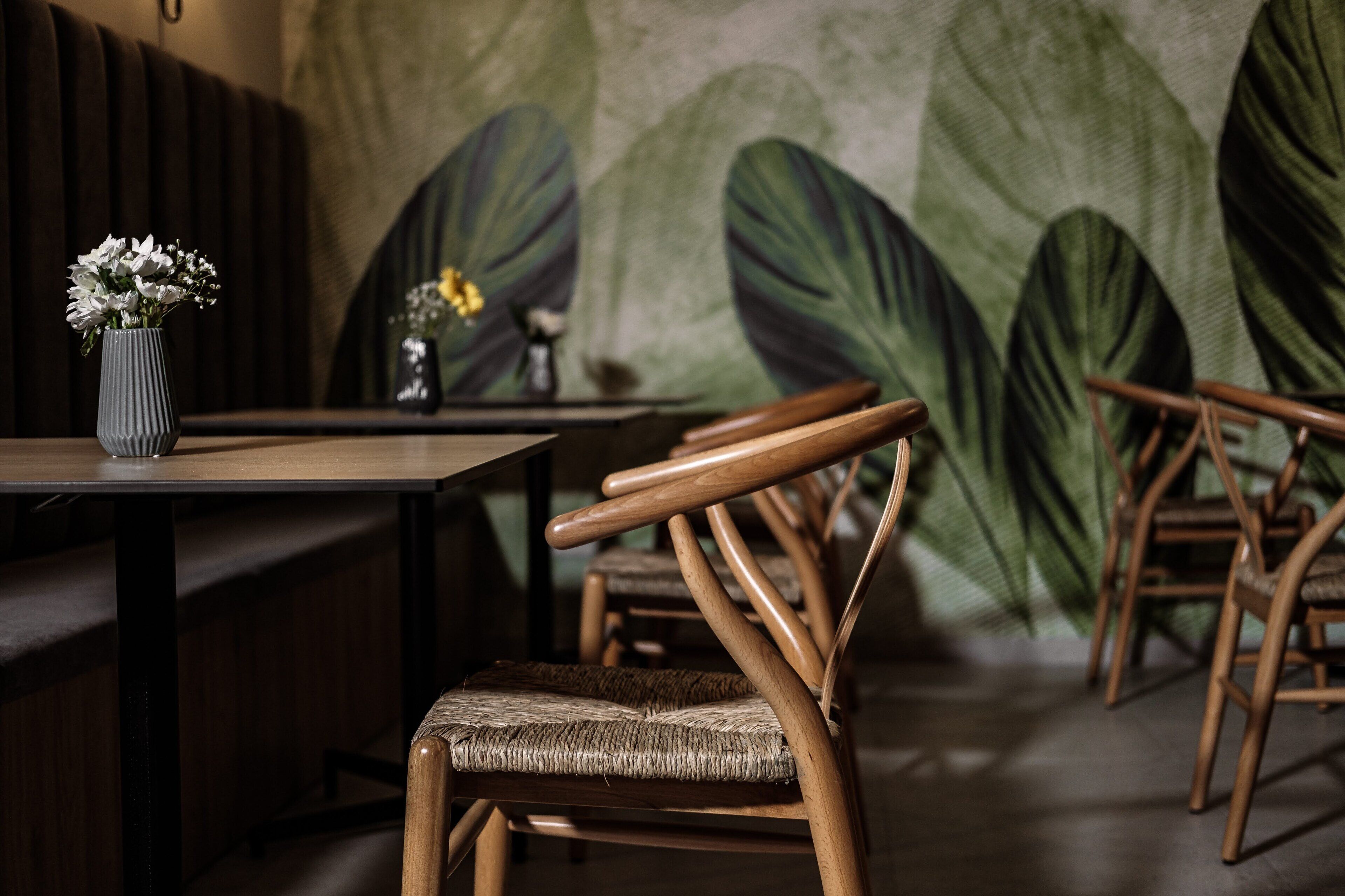 Interior de restaurante con sillas de madera, una mesa con un jarrón de flores y una pared con motivos de hojas grandes.
