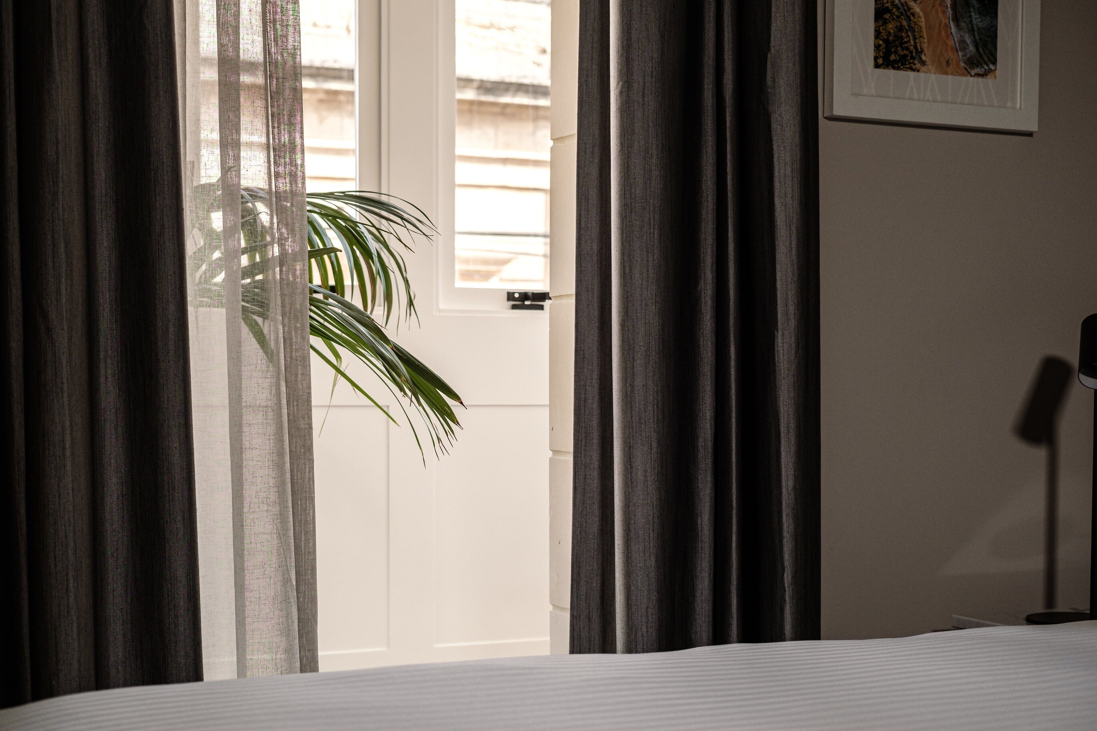 Quarto de hotel com cama, porta com cortinas, uma planta e um quadro na parede.