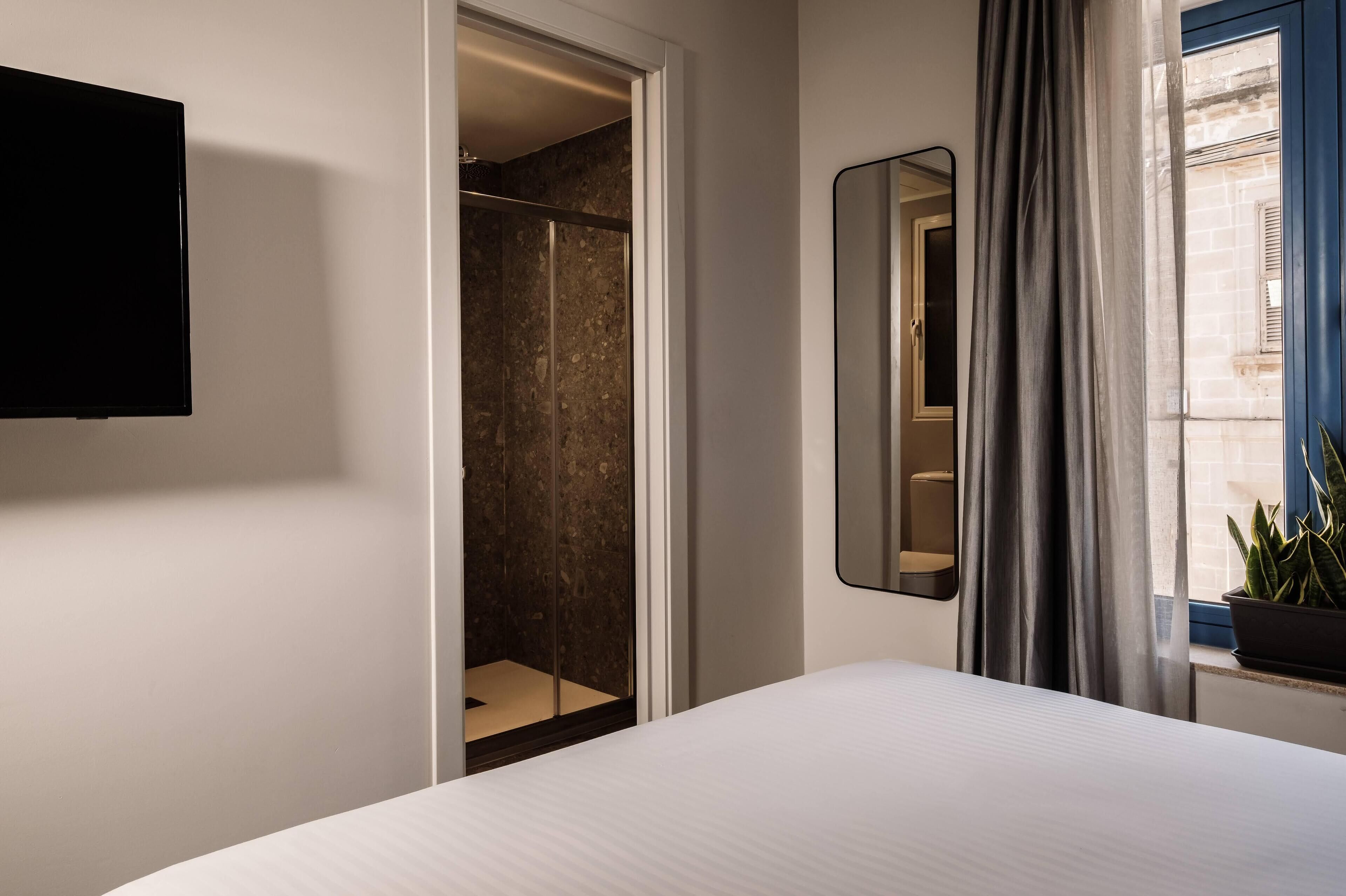 A hotel room with a bed, a TV mounted on the wall, a walk-in closet, a mirror, and a window with curtains.
