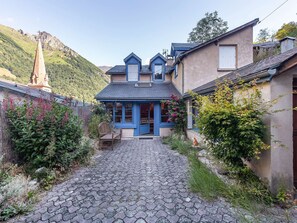 A traditional house with a stone pathway leading to the entrance, surrounded by lush greenery and a mountainous backdrop. - Charming Village House with Private Courtyard, Fireplace & WiFi (Cauterets)