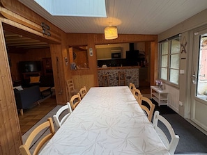 Una cabaña de madera acogedora con comedor que incluye una mesa larga y sillas, una cocina al fondo y un televisor en la sala.