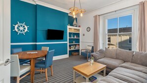 Ett modernt vardagsrum med en blå accentvägg, ett runt träbord, blå stolar, en beige soffa, en tv, en bokhylla och ett fönster med gardiner.