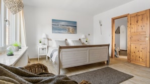 Ein Schlafzimmer mit einem Bett, Nachttischen, einem Fenster mit Pflanzen, einem Gemälde und einer Holztür.