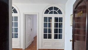Un pasillo con puertas blancas y ventanas arqueadas, una puerta de madera con el número 1502 y piso de baldosas.