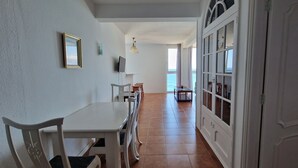 Un pasillo estrecho con un comedor que incluye una mesa y sillas blancas, un televisor montado en la pared y vistas al mar a través de un amplio ventanal.