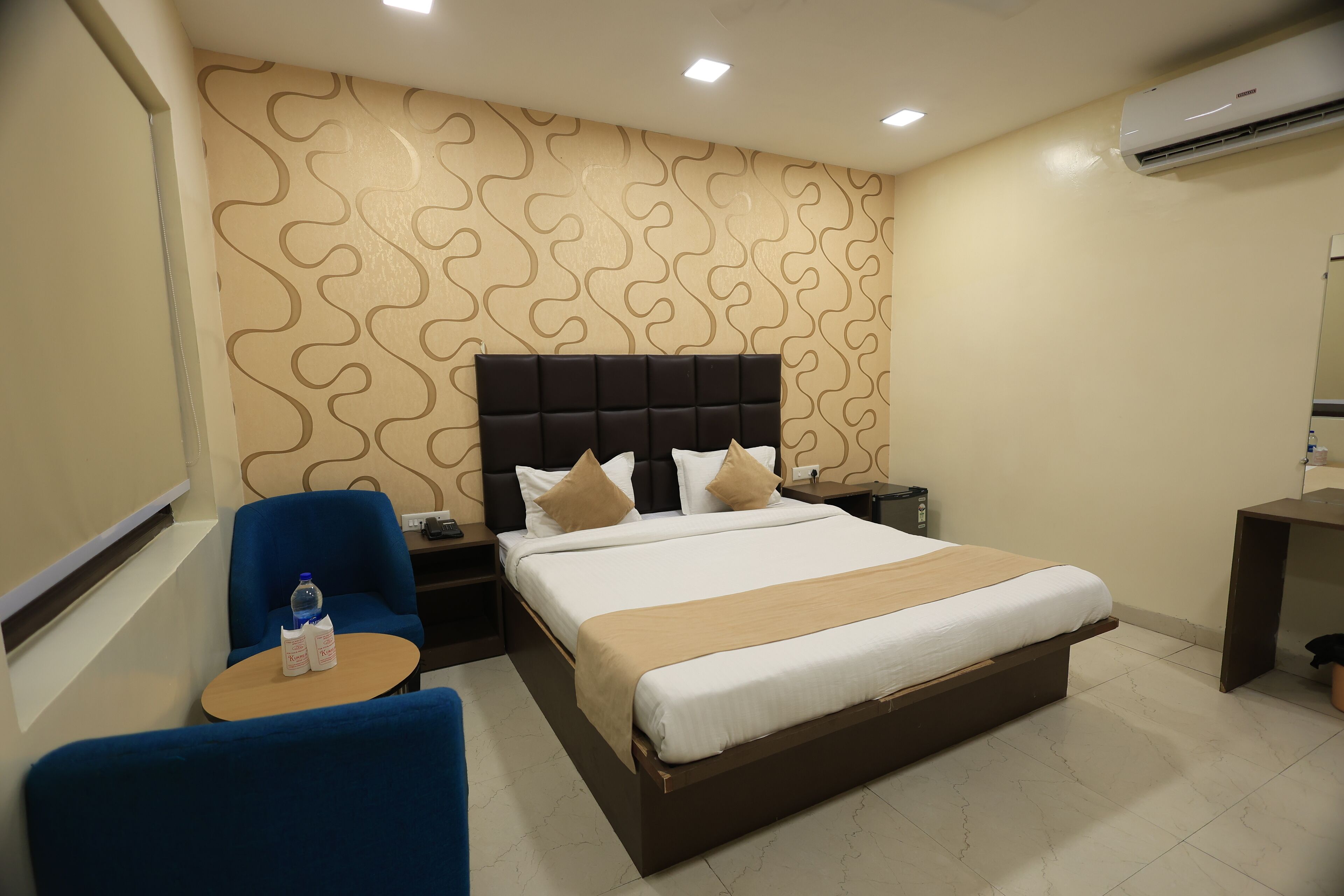 A hotel room with a large bed, a blue armchair, a small table with a bottle and glasses, and a wall-mounted air conditioner.
