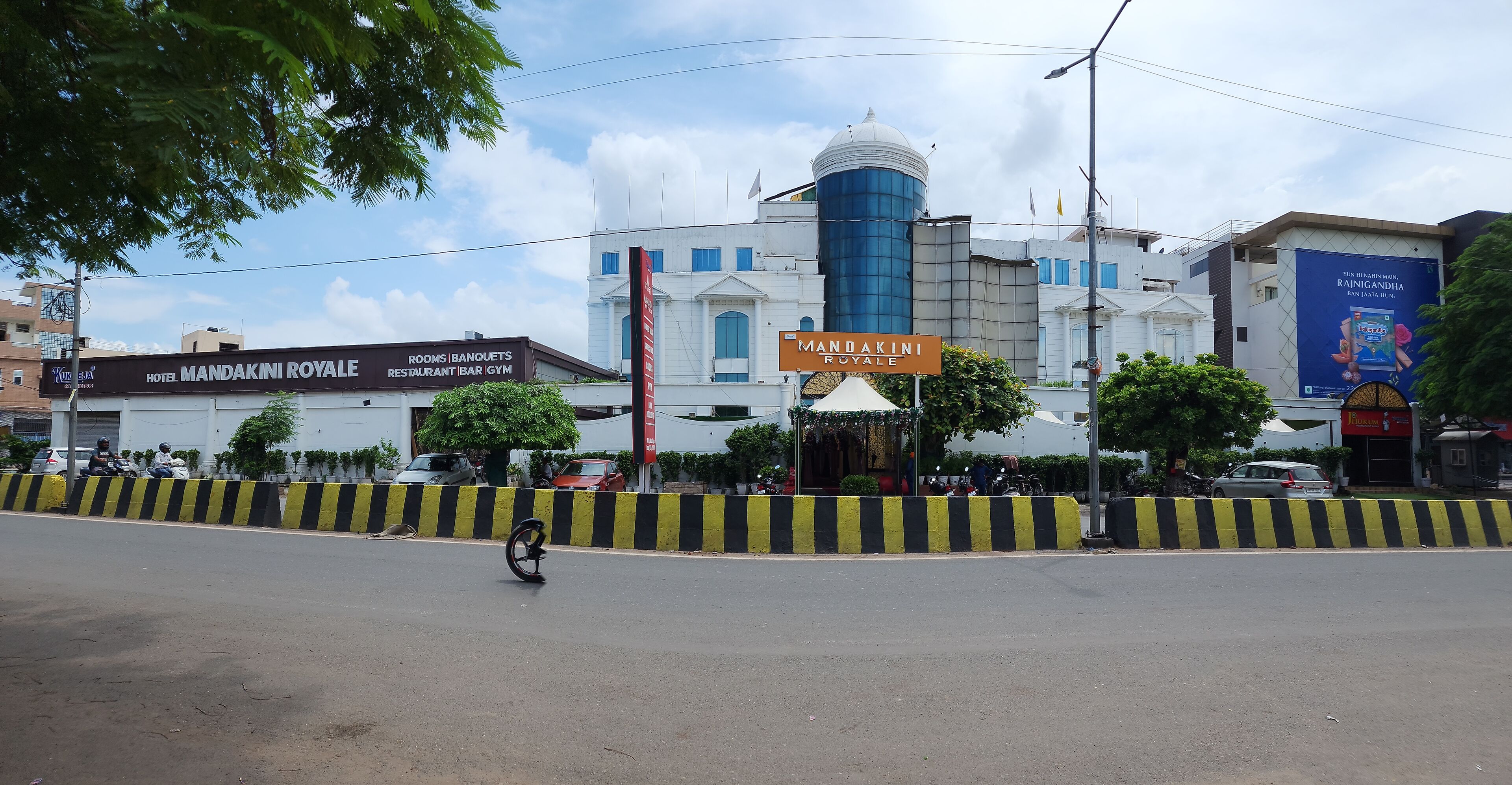 A building with a sign for 'Hotel Mandakini Royale' and 'Rooms Banquets Restaurant Bar Gym'.
