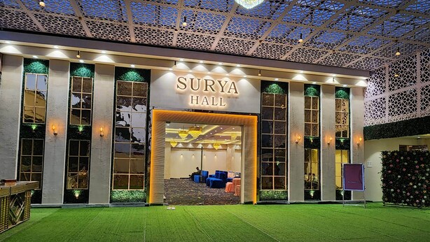 Een hotelentree met een bord waarop 'Surya Hall' staat, met een modern ontwerp, een patroonplafond en kunstgras voor.