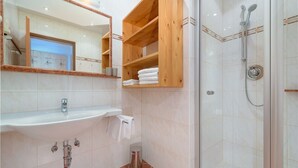 Un baño con lavamanos, espejo, estantes de madera, ducha y cubo de basura.