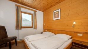 Una habitación con dos camas, paredes de madera, una ventana con cortinas, una silla y un cuadro en la pared.