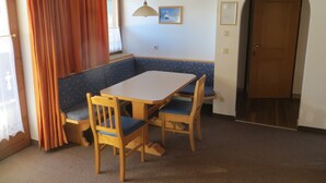 Una habitación con una mesa de madera, dos sillas, un banco, un ventilador de techo, una puerta y una ventana con cortinas.