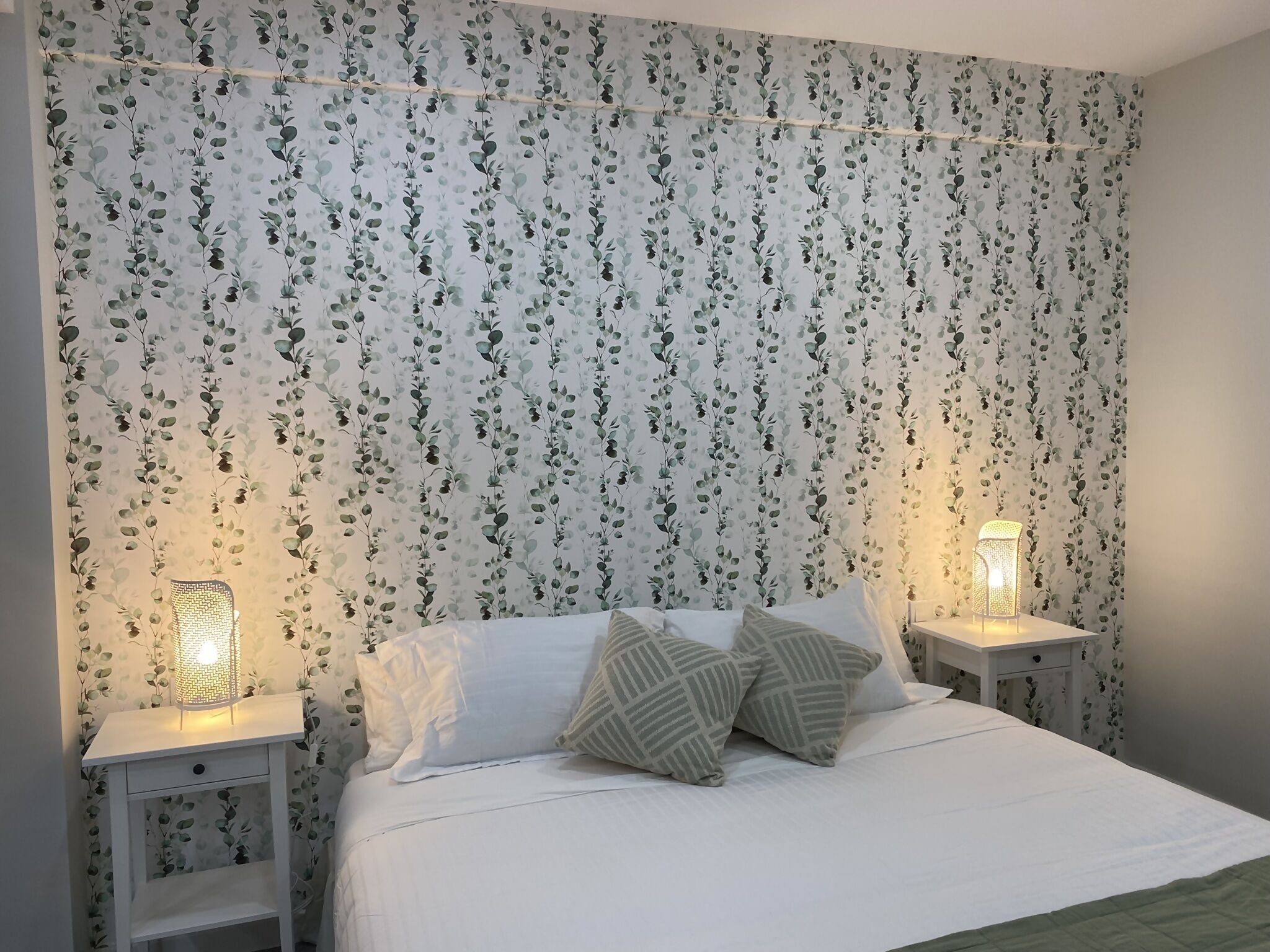 Un dormitorio con papel tapiz floral, una cama con ropa de cama blanca y almohadas estampadas, y dos mesitas de noche con lámparas.