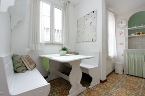 Uma cozinha pequena e acolhedora, com uma mesa branca, uma janela com cortinas, uma planta verde e um chão ladrilhado.
