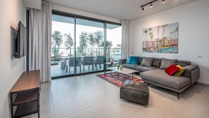 A modern living room with a grey sofa, a coffee table, and a TV on the left. There is a large window with curtains, a painting on the wall, and a balcony with outdoor furniture. - luxury garden apartment heated pool (Tiberias)