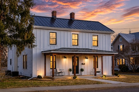 A two-story white house with a dark roof, a front porch with chairs, and a well-lit entrance.