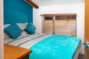 A bedroom with a bed, two pillows, a wooden headboard, a window with blinds, and a wooden nightstand. - Porthtowan Lodge eco-home, short walk to beach and coastal path. (Porthtowan)