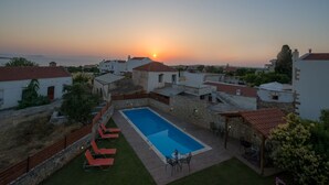 Une piscine entourée de chaises longues et d’une table, le tout bordé de murs en pierre et surmonté de toits de tuiles rouges, avec une vue sur le coucher de soleil en arrière-plan.