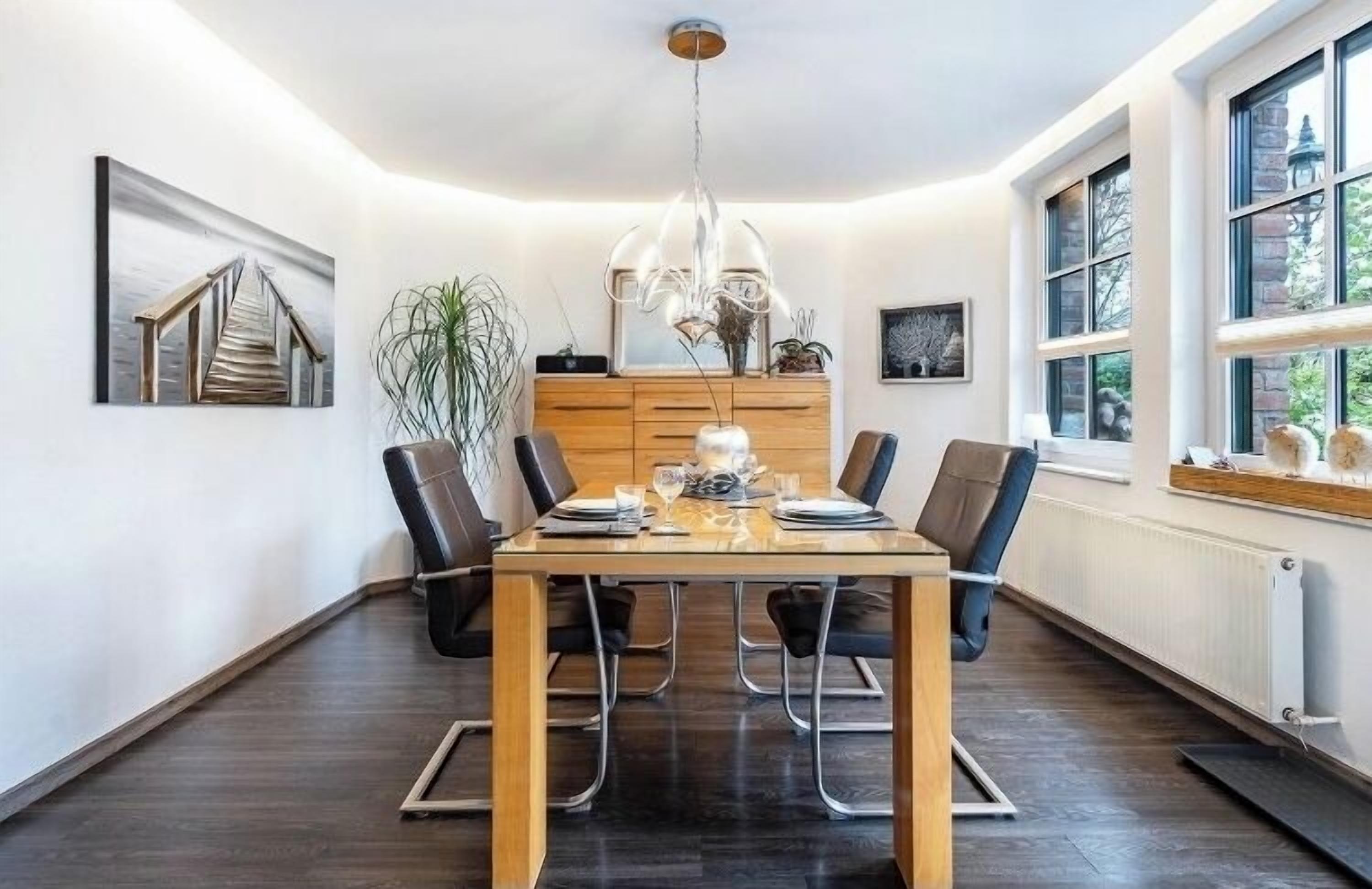 A dining area with a wooden table, black chairs, a chandelier, and a view of the outdoors through large windows.
