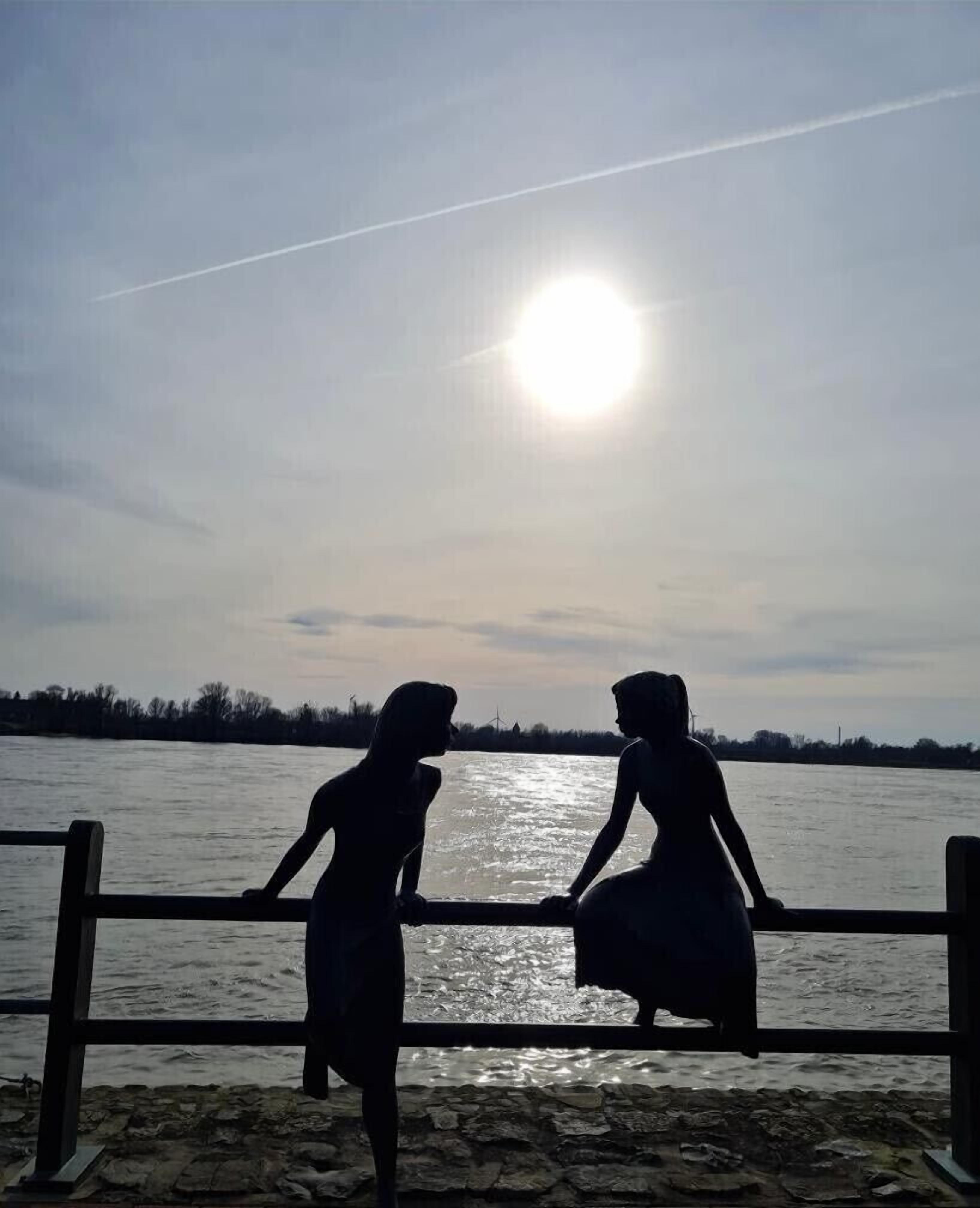 Dos siluetas de personas al lado de una barandilla, mirando un cuerpo de agua con el sol de fondo.