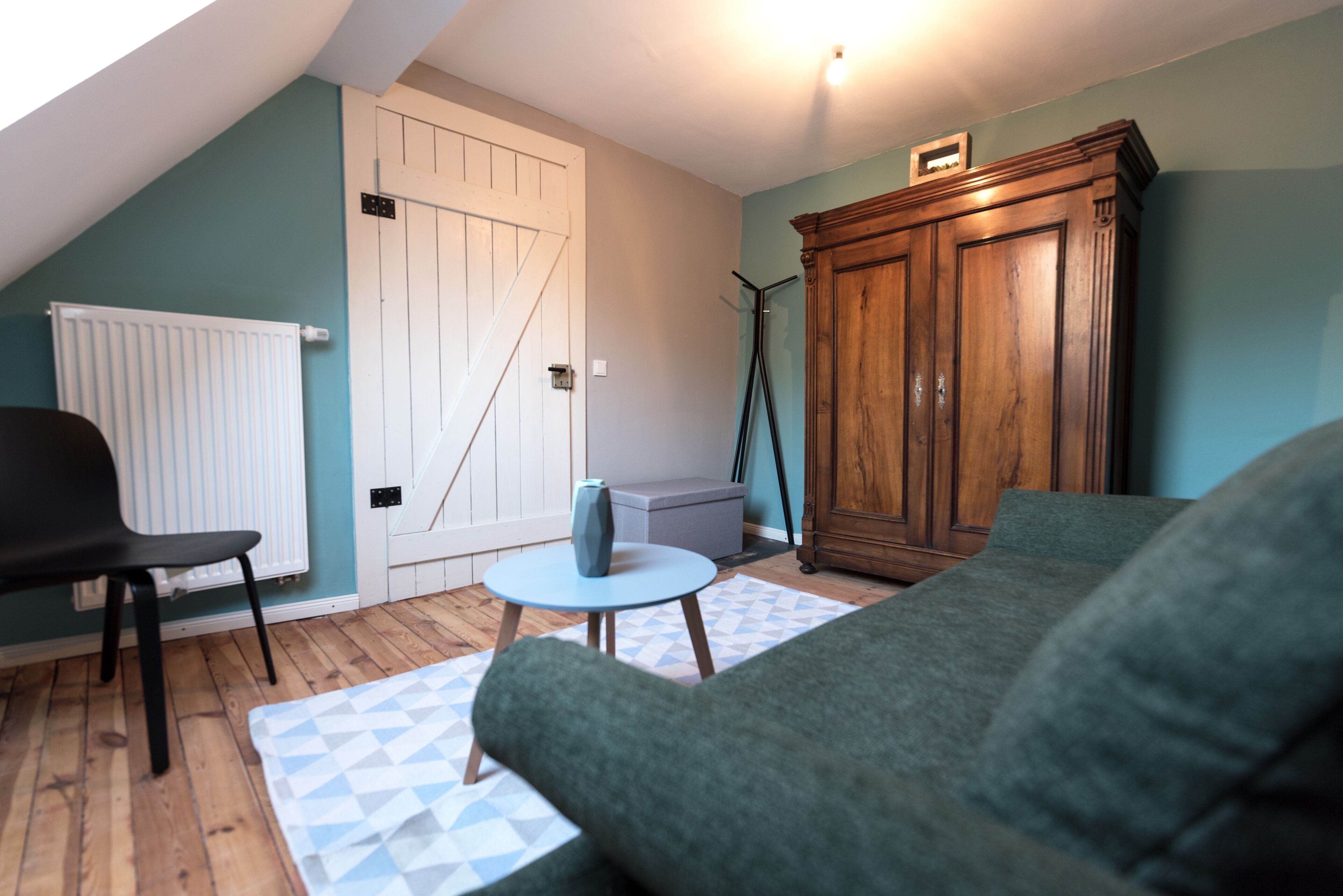 A room with a sloped ceiling, a wooden cabinet, a green sofa, a small round table, and a white radiator.