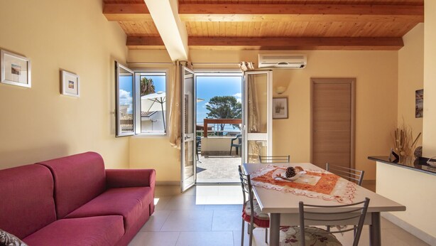 A cozy living room with a red sofa, a dining table with chairs, and a view through an open door to an outdoor patio. - Villa Tonnara 50m dal mare (Avola)