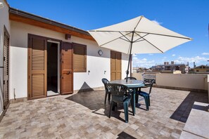 Une terrasse avec une table et des chaises, un grand parasol et des volets en bois sur un bâtiment.
