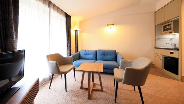 A modern hotel room with a sofa, two armchairs, a wooden coffee table, a TV, a kitchenette, and a large window with curtains. - Yutorelo Garden Kitakaruizawa (Tsumagoi)