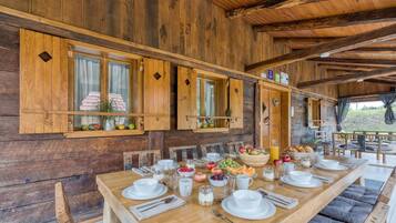 A wooden dining table set for breakfast with various fruits and beverages, wooden walls, and a view of the outdoors.