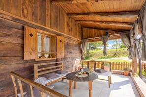 Uma cabana de madeira com varanda, um banco, uma mesa com uma tigela e um jarro, e vista para uma paisagem verde.