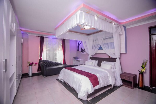 A bedroom with a four-poster bed, a bedside table, a nightstand, a wardrobe, and a window with curtains. - Lovely Garden Guest Home (Kisumu)