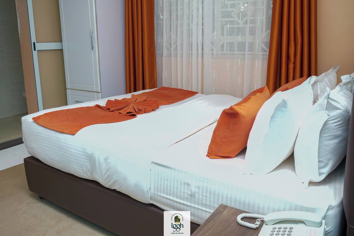 Habitación de hotel con una cama, almohadones naranjas, una manta blanca, una mesita de noche de madera con un teléfono y una ventana con cortinas naranjas.