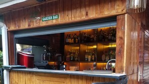 Un coin bar en bois, agrémenté d’une enseigne indiquant « Garden Bar », et équipé d’une machine à café et d’étagères remplies de bouteilles. On y trouve deux chaises noires avec des assises en osier tressé.