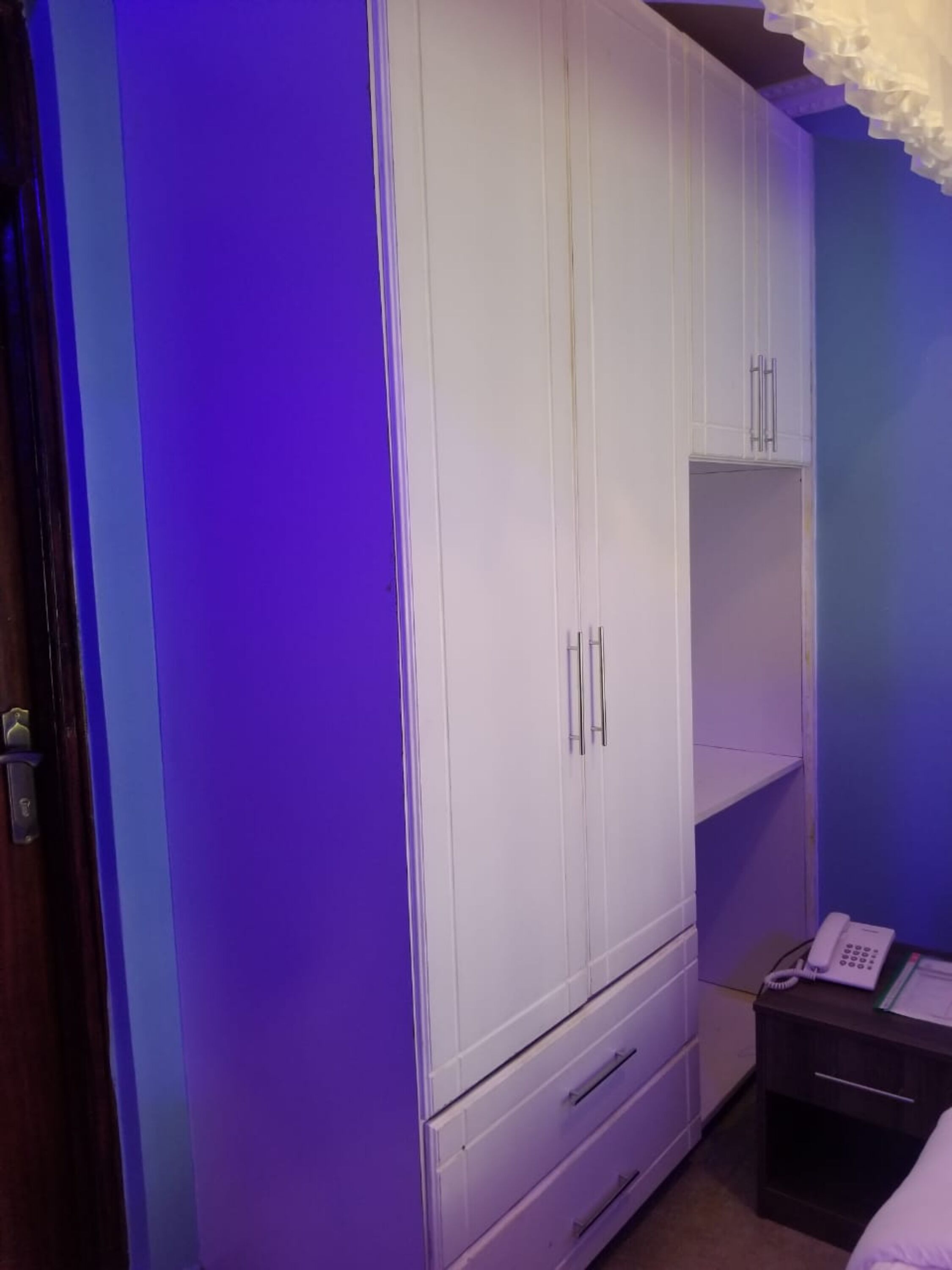 Een slaapkamer met een witte kast, een nachtkastje met een telefoon, en een paarse muur.