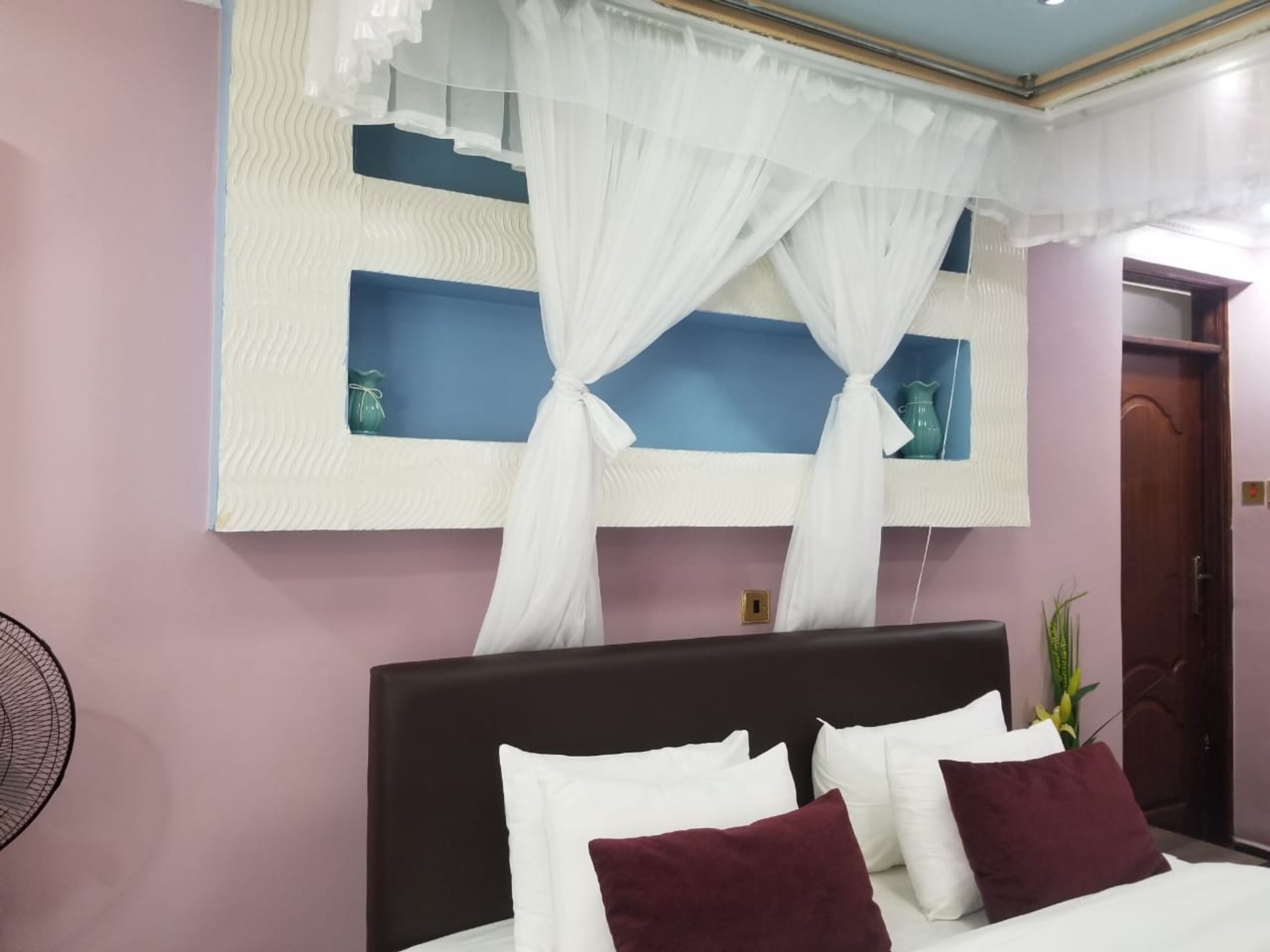 A bedroom with a bed, white curtains, a wall-mounted shelf, and a door.