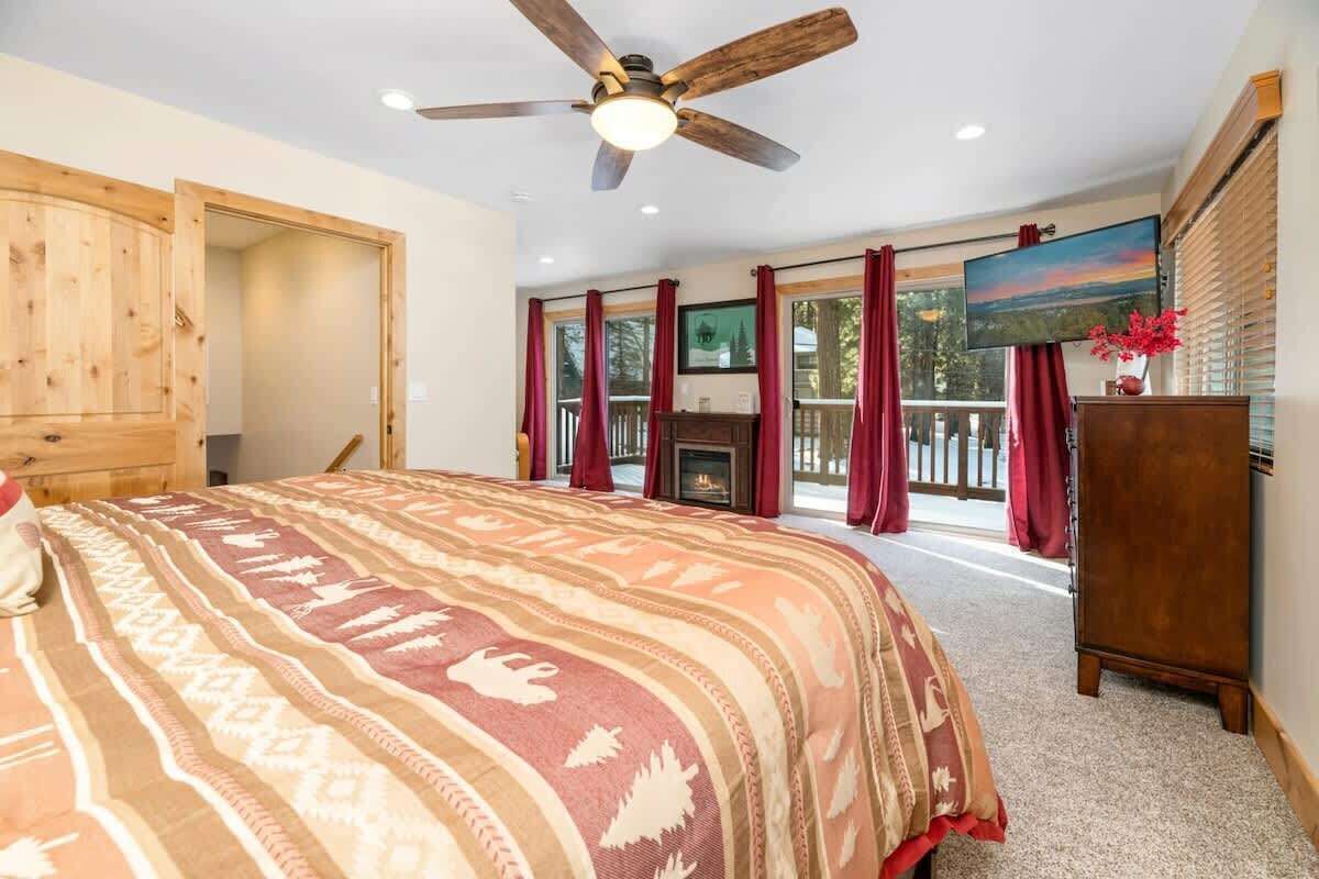 A bedroom with a large bed, a ceiling fan, a fireplace, and a view of the outdoors through large windows.
