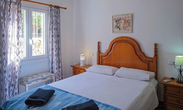 A bedroom with a wooden bed, a bedside table, a lamp, and a window with curtains.