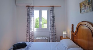 Ein Schlafzimmer mit einem Bett, einem Nachttisch mit Lampe, einem Fenster mit Vorhängen und einer Klimaanlage.