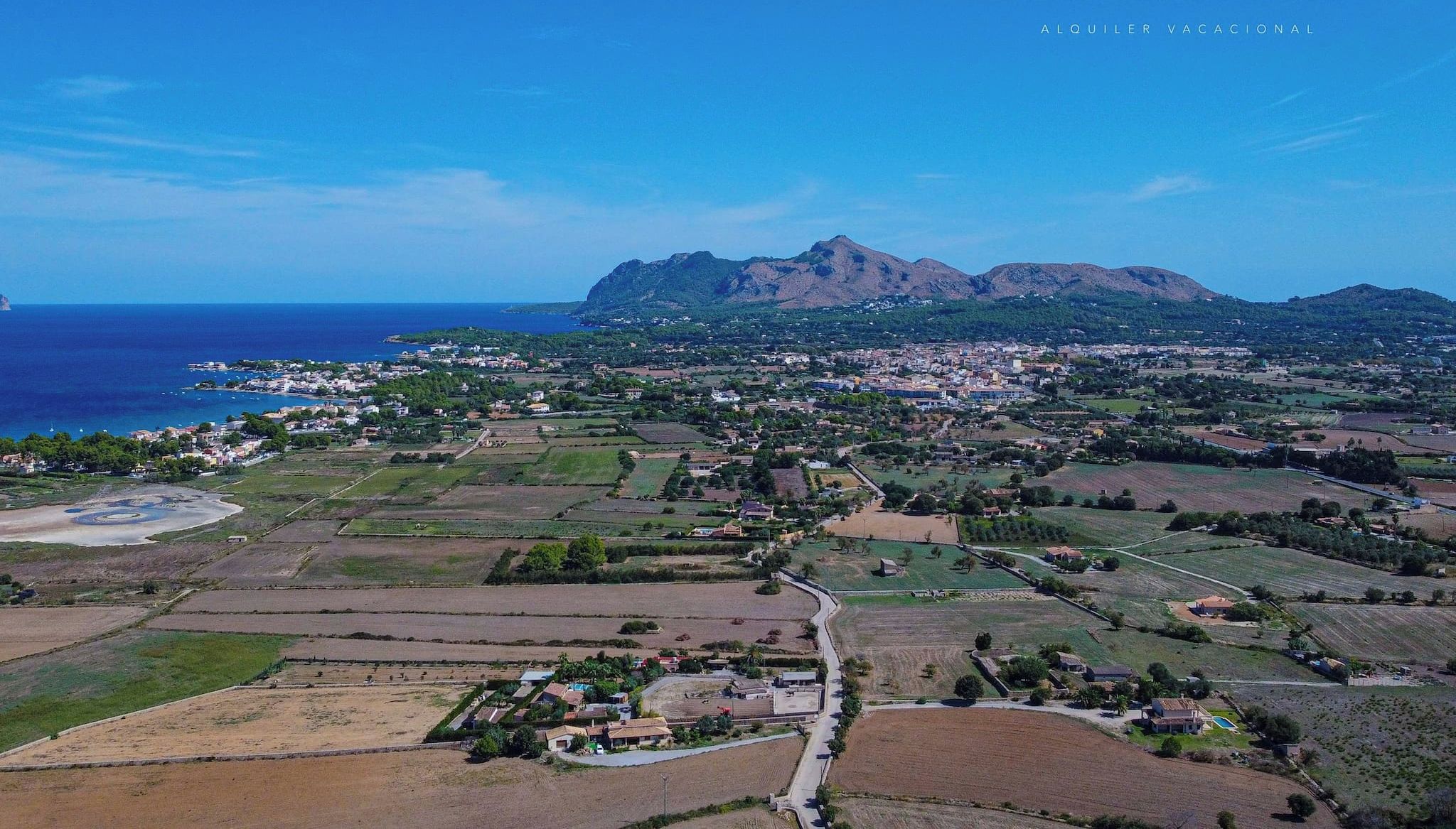 Vue aérienne d’une ville côtière, avec des champs, une plage et des montagnes en arrière-plan.