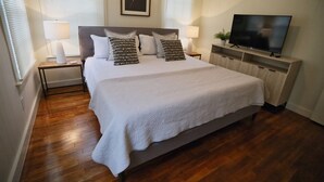 Ein ordentlich eingerichtetes Schlafzimmer mit einem großen Bett, zwei Nachttischen mit Lampen, einem Flachbildfernseher auf einem Holzständer und einem gerahmten Kunstwerk über dem Bett.