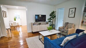 Ein modernes Wohnzimmer mit einem blauen Sofa, einem weißen Couchtisch, einem Fernseher auf einem Holzschrank und einem Essbereich mit Tisch und Stühlen in Weiß.