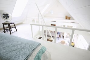 A high-ceilinged room with a bed, a standing fan, and a staircase leading to an upper level.