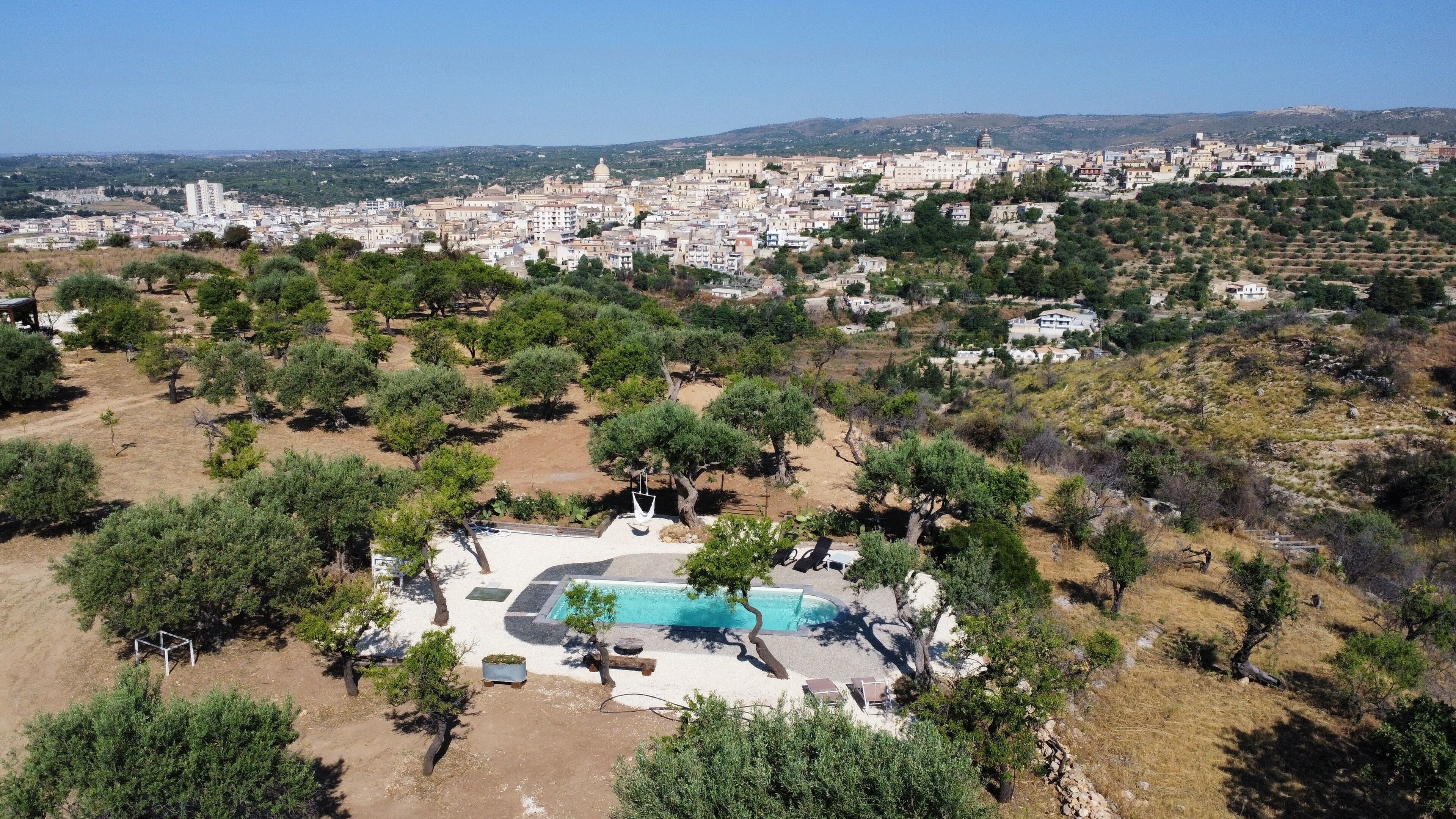 An aerial view of a residential area with a swimming pool surrounded by trees and a clear view of a distant cityscape.