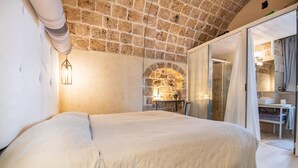 Un dormitorio con techo de piedra, una cama con colcha blanca, una lámpara fijada a la pared y una puerta de vidrio que da a un baño.
