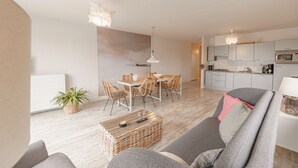 Un salon moderne avec un canapé gris, une table à manger entourée de chaises en osier, une cuisine avec des meubles blancs et un grand tableau abstrait accroché au mur.
