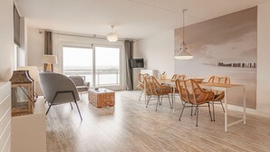 Un salon moderne avec un canapé gris, une table à manger entourée de chaises en osier et une grande fenêtre donnant sur la mer.