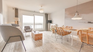Un salon moderne comprenant un canapé, une table à manger avec des chaises en osier, un téléviseur et une grande fenêtre offrant une belle vue.