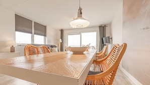 Un espace repas avec une longue table et des chaises en osier, un luminaire suspendu et une vue sur un balcon à travers de grandes fenêtres.