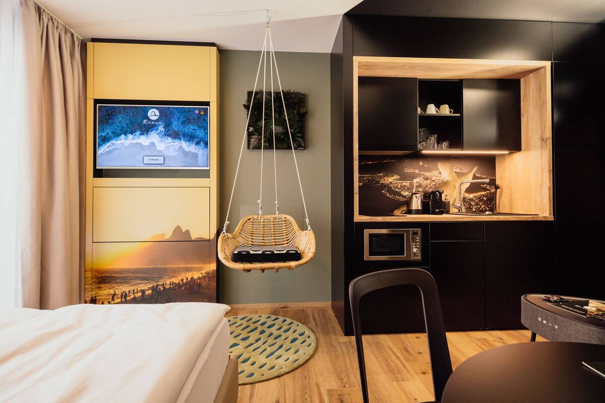 A modern hotel room with a bed, a hanging chair, a dining table, and a kitchen area with a wooden countertop.