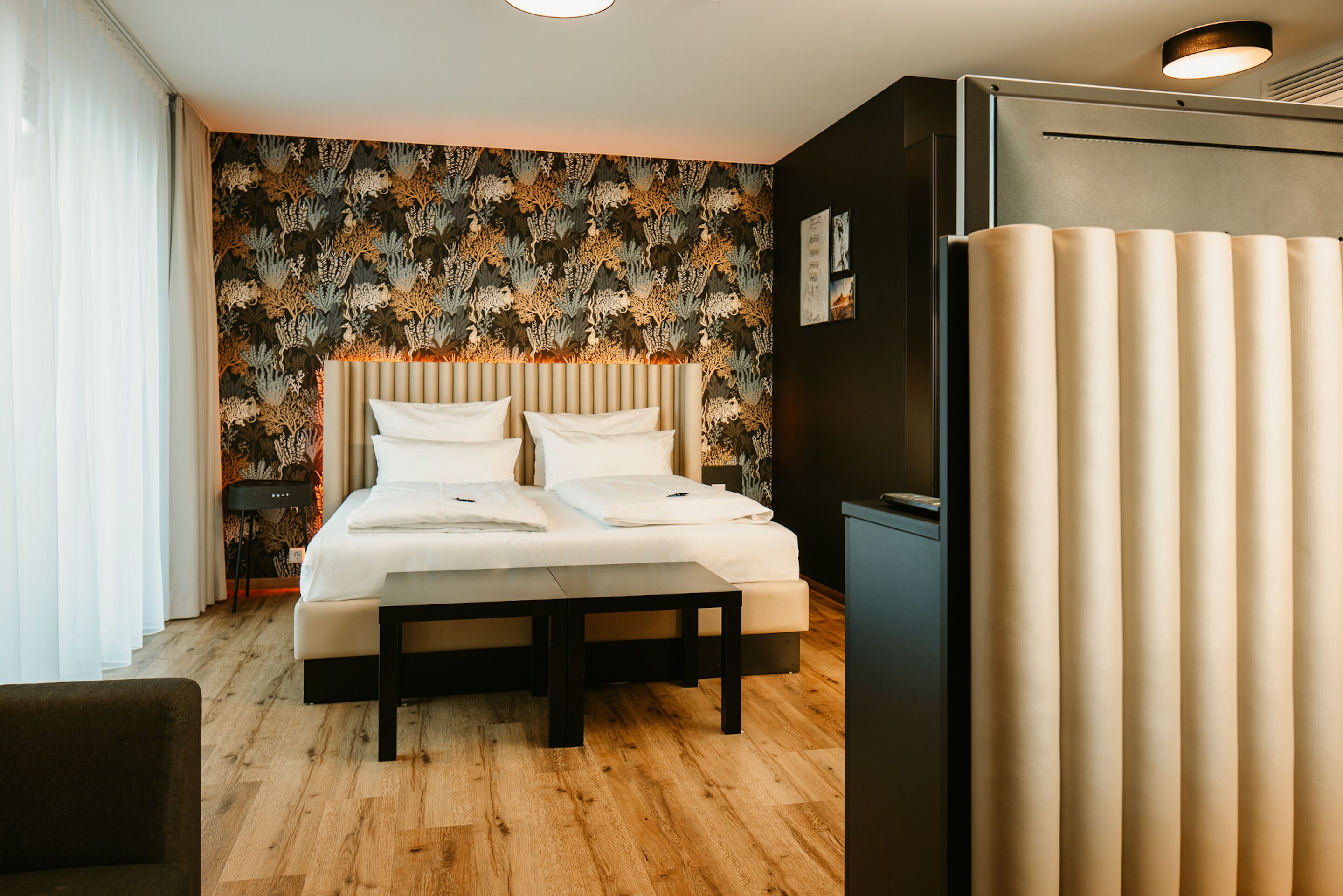 Ein modernes Hotelzimmer mit einem großen Bett, einem Nachttisch, einer Ablagefläche und einer Wand mit floralem Dekor.